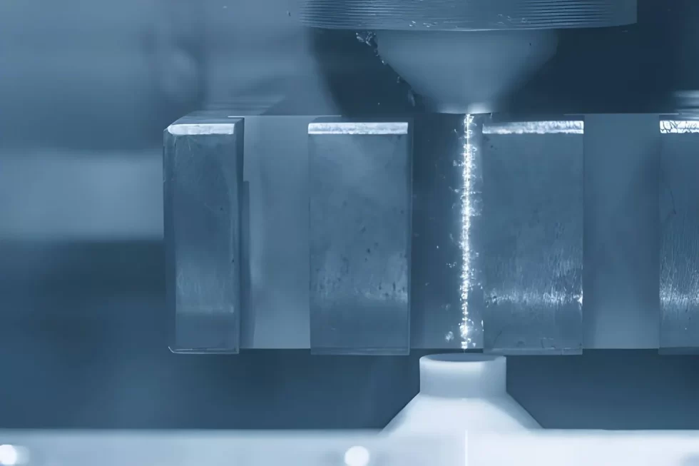 The image shows a close-up of a CNC-controlled wire EDM machine during machining, with a blue-toned appearance.