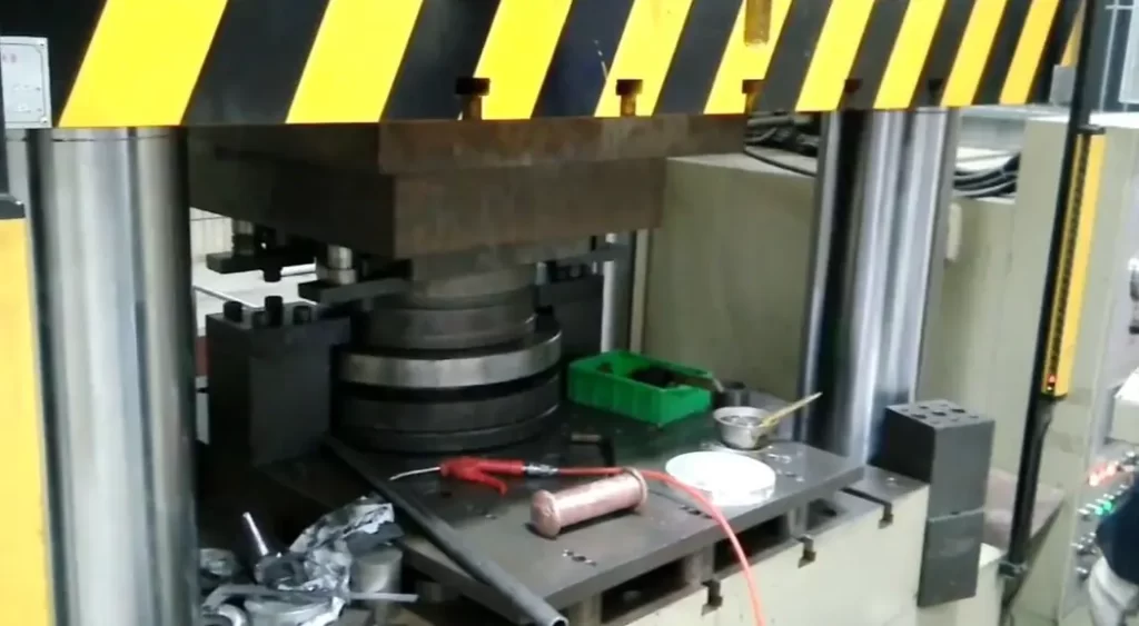 Industrial cold forging process showing a press machine with yellow and black safety striping and metal workpieces on the platform 