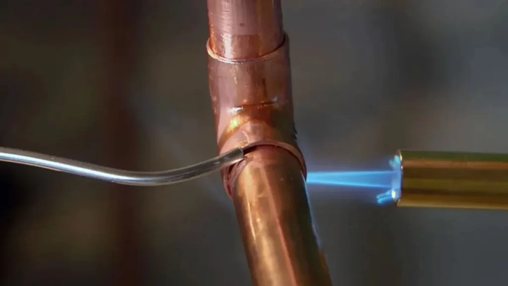 A close-up of a worker using a torch to solder two copper pipes together.